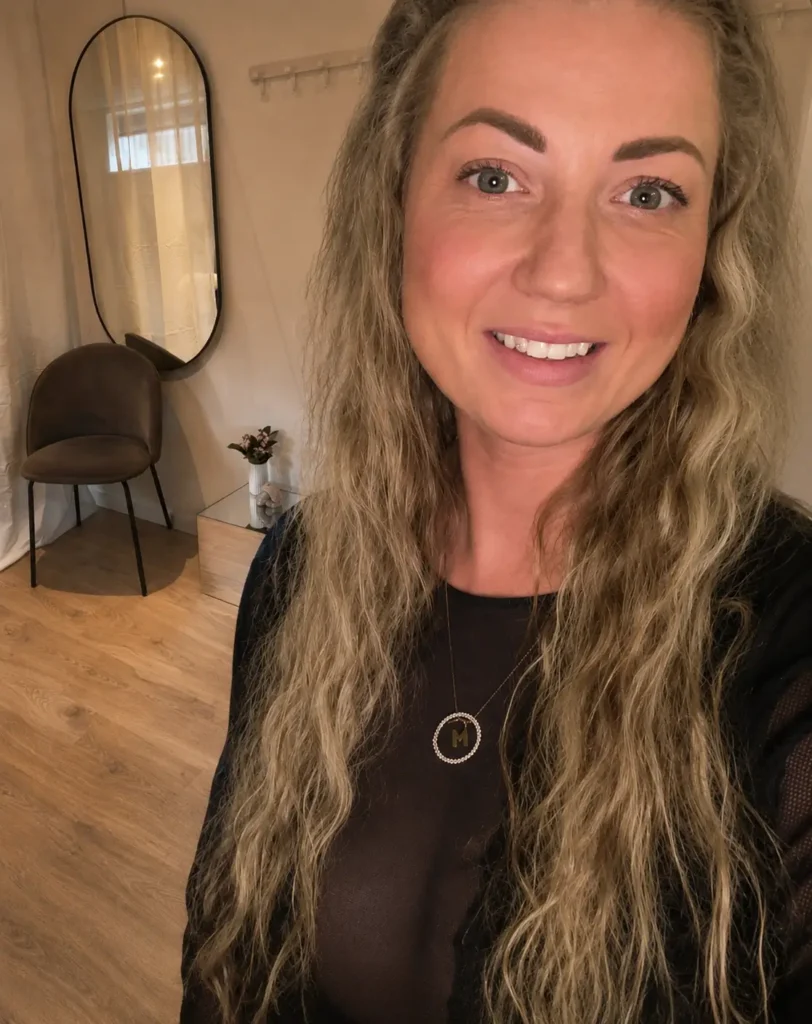 Selfie af BeautybyRosens ejer, der smiler og kigger direkte ind i kameraet, med dele af skønhedssalonen synlig i baggrunden.